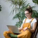 Woman gardener sitting on wooden chair in greenhouse, using laptop after work, talks with her friend about coronavirus and stay home during online video call. Working home